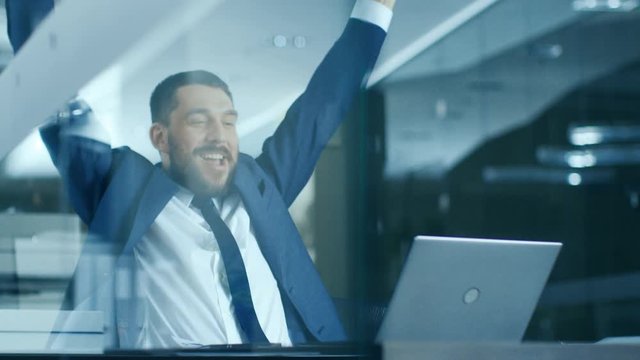 In the Office Businessman Sitting at the Desk Using Laptop Finishes Project and Wins Big. Makes Successful Gestures Raises Arms in Celebration. Shot on RED EPIC-W 8K Helium Cinema Camera.