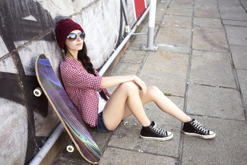 Brunette teenage girl in hipster outfit (jeans shorts, keds, plaid shirt, hat) with a skateboard at the park outdoors