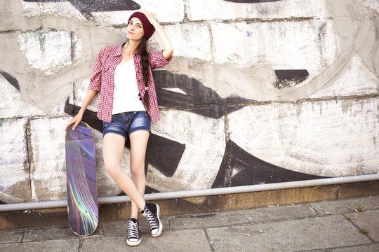 Brunette teenage girl in hipster outfit (jeans shorts, keds, plaid shirt, hat) with a skateboard at the park outdoors