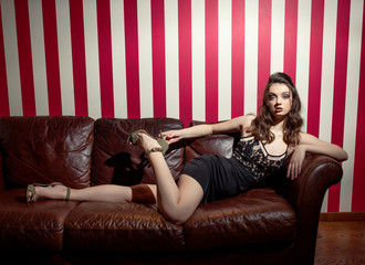 A young girl in a tight black dress sits on the couch. Leather sofa - obrazy, fototapety, plakaty