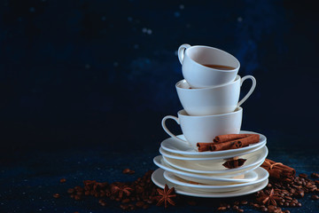 Obraz na płótnie Canvas Header with a stack of balancing white coffee cups and tea saucers, coffee beans and cinnamon on a dark blue background. Dark food photography with copy space.
