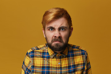 Fototapeta premium Young bearded male feels angry and mad about some problems and people. Wearing shirt, blue eyes and dyed blond hair. Isolated over yellow background.