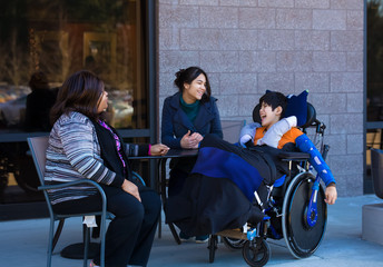 Disabled boy in wheelchair at table outdoors talking with caregivers