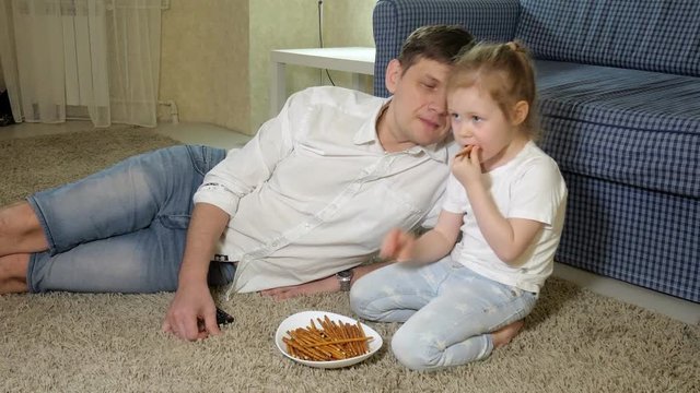 man and daughter watching television, sitting on the floor eating snacks