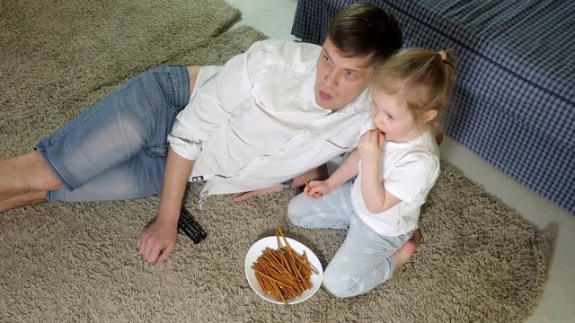 man and daughter watching television, sitting on the floor eating snacks