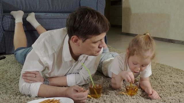 man and daughter watching television, sitting on the floor drink juice