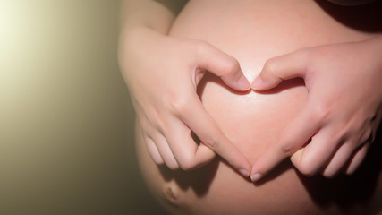 woman holding her hands in a heart shape on her baby bump pregnant belly with fingers on black background