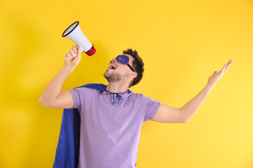 Young man in superhero costume with megaphone on color background