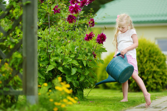 Cute little girl watering flowers in the garden at summer day. Child using garden hose on sunny day