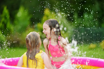 Adorable little girls playing in inflatable baby pool. Happy kids splashing in colorful garden play center on hot summer day. © MNStudio