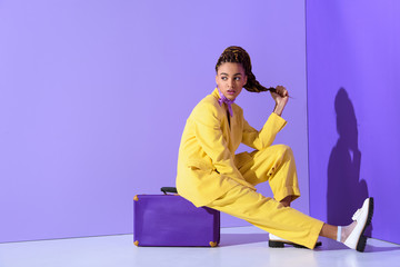african american girl posing in yellow suit sitting on purple suitcase, on trendy ultra violet...