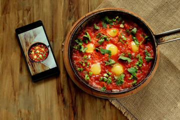 Fried eggs in tomato sauce in a cast iron pan and a smartphone with the photo on a wooden table