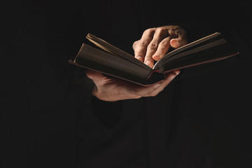 Priest with old Bible on black background, closeup