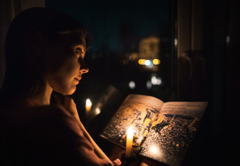 A beautiful young girl in a shirt reading a book at the window-sill by the light of a burning...
