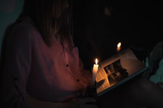 A Beautiful Young Girl In A Shirt Reading A Book At The Window-sill By The Light Of A Burning Candle At Night