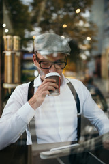 Fashionable retro dressed man with cap, suspenders and eyeglasses sitting in cafe bar and reading a newspapers. View through window glass.