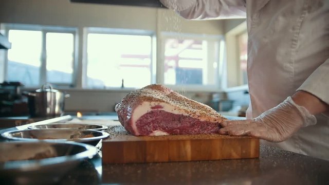Butcher adding spices into large piece of fresh raw meat lying on a wooden board in a commercial kitchen, slow motion