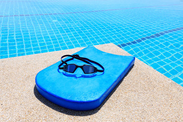 Black sport goggle on blue kick board beside clear swimming pool 