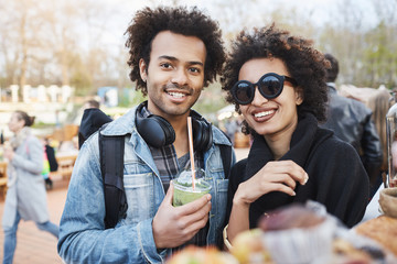 Portrait of happy cute dark-skinned couple with afro hairstyle, strolling on food festival, tasting...