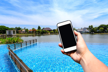 Hand holding blank screen smartphone with beautiful sky and outdoor swimming pool and lake with copy space.