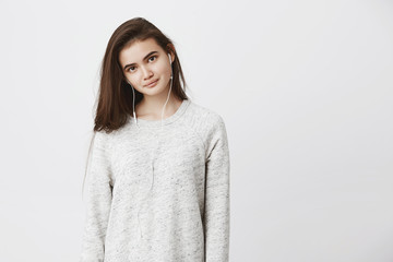Fototapeta premium Portrait of beautiful tender young adult woman, standing straight over white background while smiling and wearing earphones. Cute girl learns foreign language listening audio course she uploaded