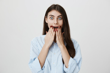 Indoor portrait of amazed and surprised stylish woman standing with opened mouth and hands on face, staring at camera while standing over gray background. Girl received amazing present from her lover