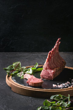 Grilled sliced rack of lamb with yogurt mint sauce served with green salad young beetroot leaves and pink salt on round wooden slate board over dark black table background. Copy space