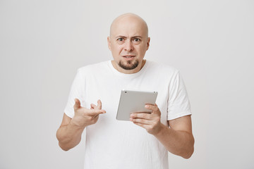 Indoor portrait of irritated upset bald caucasian male holding tablet and pointing at it, while frowning and being puzzled and unsatisfied with what he read, standing against gray background.