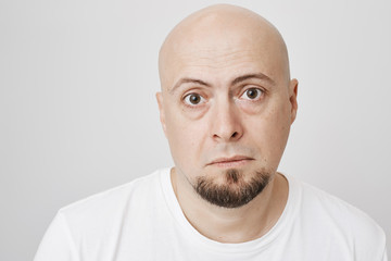 Close-up portrait of upset tired unhappy bearded bald man, looking tired, standing against gray background. Exhausted miserable guy having bad day, begging brother to cover him at work