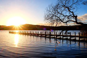 Fototapeta premium very long wooden jetty, jutting out into a calm blue wooden lake with mountains sunsetting in background