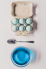 top view of glass with blue paint, spoon and chicken eggs in tray, easter concept