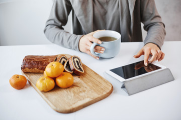 Side angle shot of female hands with manicure touching digital tablet. Student having breakfast before going to university, drinking cup of tea and eating tangerines with rolled cake she baked herself