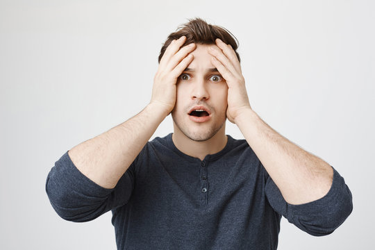 Stressed frustrated dark-haired young male with stubble dressed in casual clothes holds hands on head, keeps mouth opened in despair, doesn`t know what to do in stressful situation, feels lost