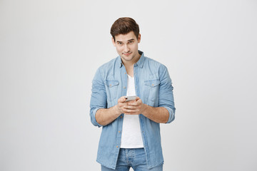 Young attractive male model holding smartphone while looking at camera, isolated over white background. Man was writing note in gadget, but he got distracted because someone called his name