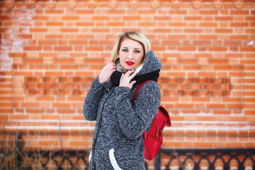 A beautiful hipster girl in a gray coat smiles and looks at the camera on a red background outdoors. Portrait of a modern girl with red lips against a brick wall background.