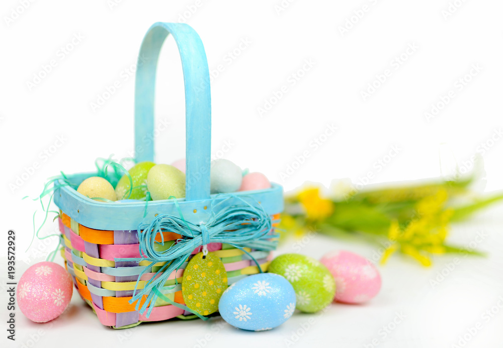 Wall mural A colorful Easter basket full of sparkly eggs in blue, pink, yellow and green with copy space on a bright white background. Shallow depth of field with yellow flowers in the distance.