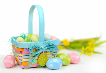 A colorful Easter basket full of sparkly eggs in blue, pink, yellow and green with copy space on a bright white background. Shallow depth of field with yellow flowers in the distance.