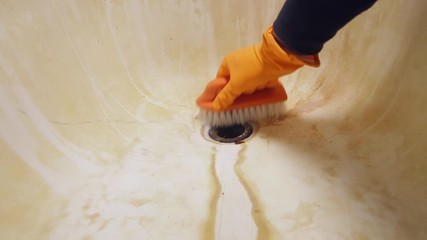 A man's hand in orange rubber gloves cleans the bathroom sink with a scrub brush. General house cleaning.