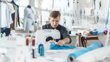 Fototapeta premium Male fashion, designer, / Dressmaker Working on a Sewing Machine in His Sunny Studio. Various Sewing items and Fabrics Laying Around Him, Visible Sketches Pinned to the Wall.