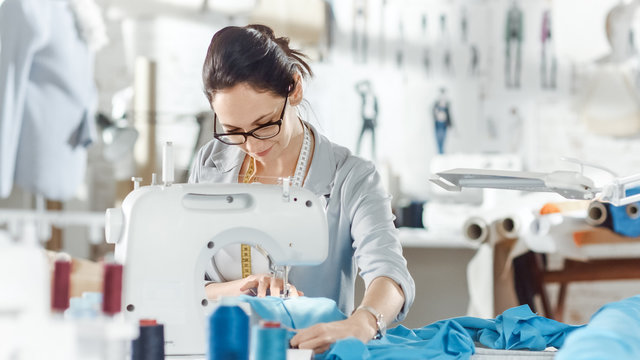 Female fashion, designer, / Seamstress/ Dressmaker Working on a Sewing Machine in Her Sunny Studio. Various Sewing items and Fabrics Laying Around Her, Visible Sketches Pinned to the Wall.