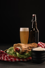 tasty french hot dog on plate with bottle and glass of beer isolated on black