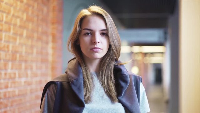 Calm and beautiful young woman looking at camera and blinking while standing in a building corridor with brick walls. Handheld slow motion medium shot