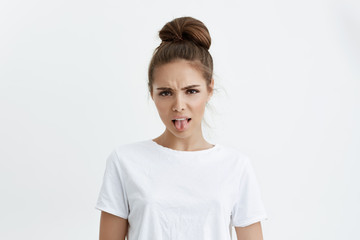 Obraz premium Studio portret nieszczęśliwej kobiety rasy kaukaskiej wyrażającej niechęć lub wstręt, pokazując język i marszcząc brwi, będąc rozczarowaną i stojącą na szarym tle. Dziewczyna nienawidzi filmów o miłości