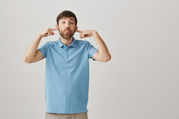 Guy not going to listen to excuses. Portrait of carefree attractive bearded guy covering ears with index fingers and sticking out tongue, being childish while standing over gray background.