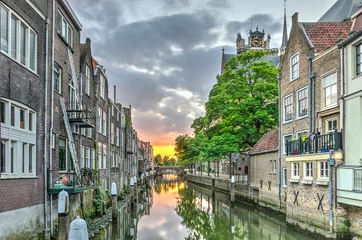Fotobehang A canal in the old medieval town centre of Dordrecht, the Netherlands, with houses on both sides and an orange red sunset in the distance © Frans