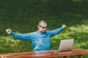 Young man businessman or student in casual blue shirt, glasses relaxing, sitting at table with laptop, mobile phone in city park stretching, spreading hands, working outdoors. Mobile Office concept.