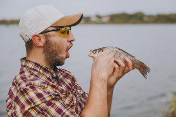 Close up Young unshaven smiling man in checkered shirt, cap and sunglasses caught a fish and opened...