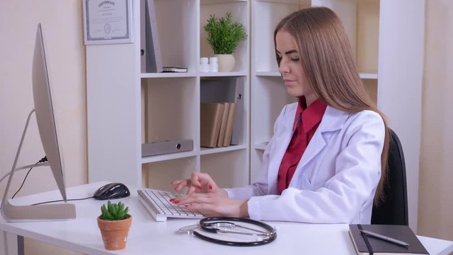 attractive doctor typing on laptop filling medical history or chatting online or research something in internet. happy beautiful therapist wearing in medical coat sitting in the office with white