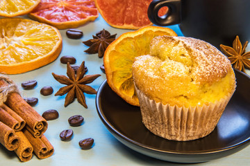 Cupcake sprinkled with sugar powder on a black plate next to a cup of coffee with cinnamon beans and chopped orange