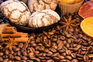 On the scattered beans of coffee there is a plate with chocolate cookies next to the sticks of cinnamon cake, green twig and sliceed orang